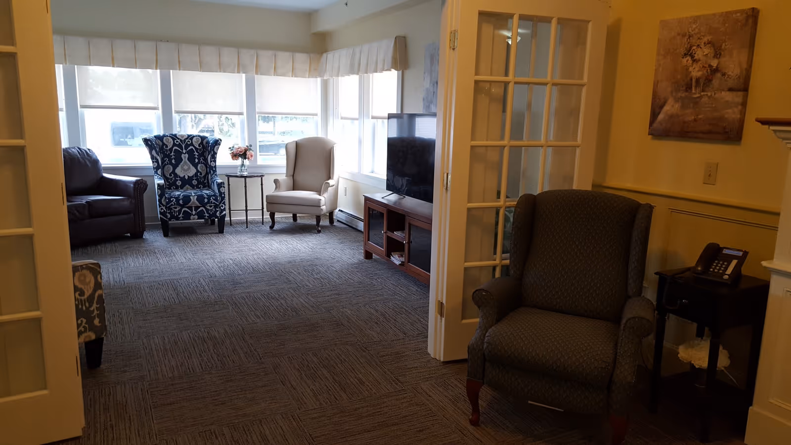 Sunlit communal sitting room with multiple upholstered armchairs, a TV on a console, side tables, and large windows.