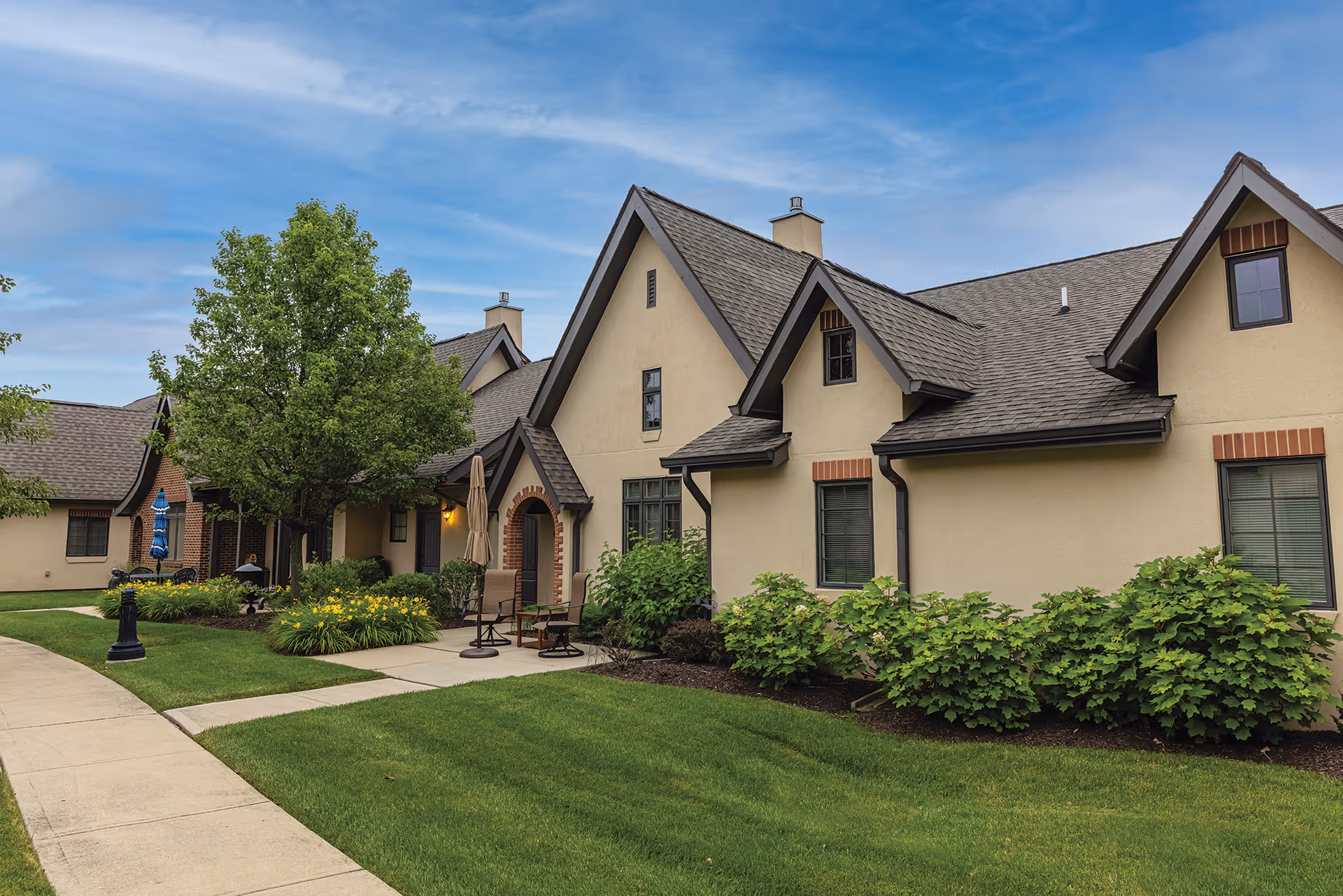 Exterior view of a senior living facility named The Stratford, showing beige buildings with dark roofs, windows, and small patios with chairs and umbrellas. The area is landscaped with green grass, bushes, flowers, and a tree under a blue sky with some clouds.