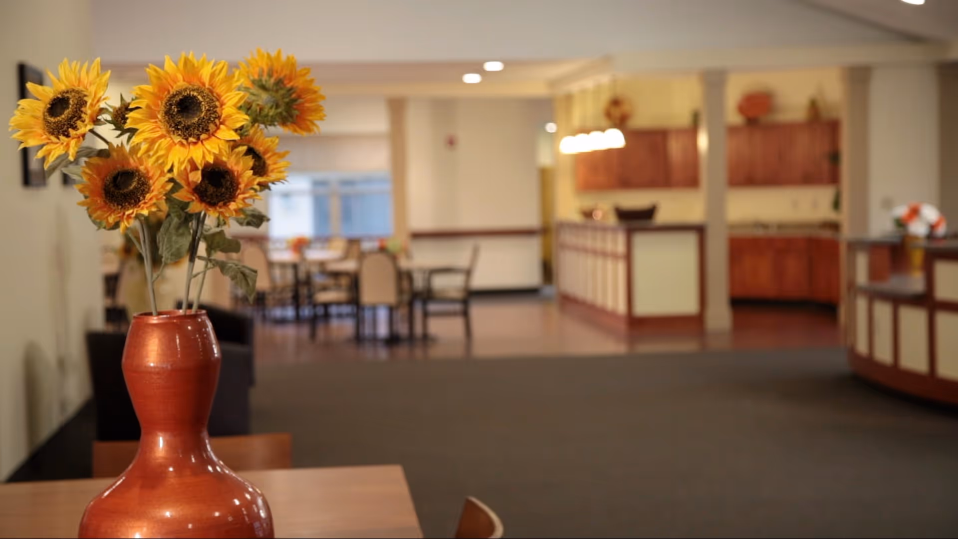 A vase of sunflowers on a table in the foreground with a dining/common area, tables, chairs, and a service counter in the background.