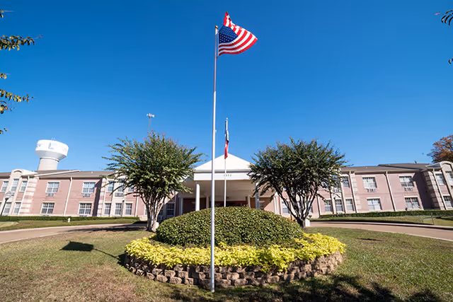 Front exterior view of The Eastman Senior Living facility with a circular landscaped area featuring bushes and yellow flowers, two flagpoles with the American flag and another flag, and a clear blue sky.