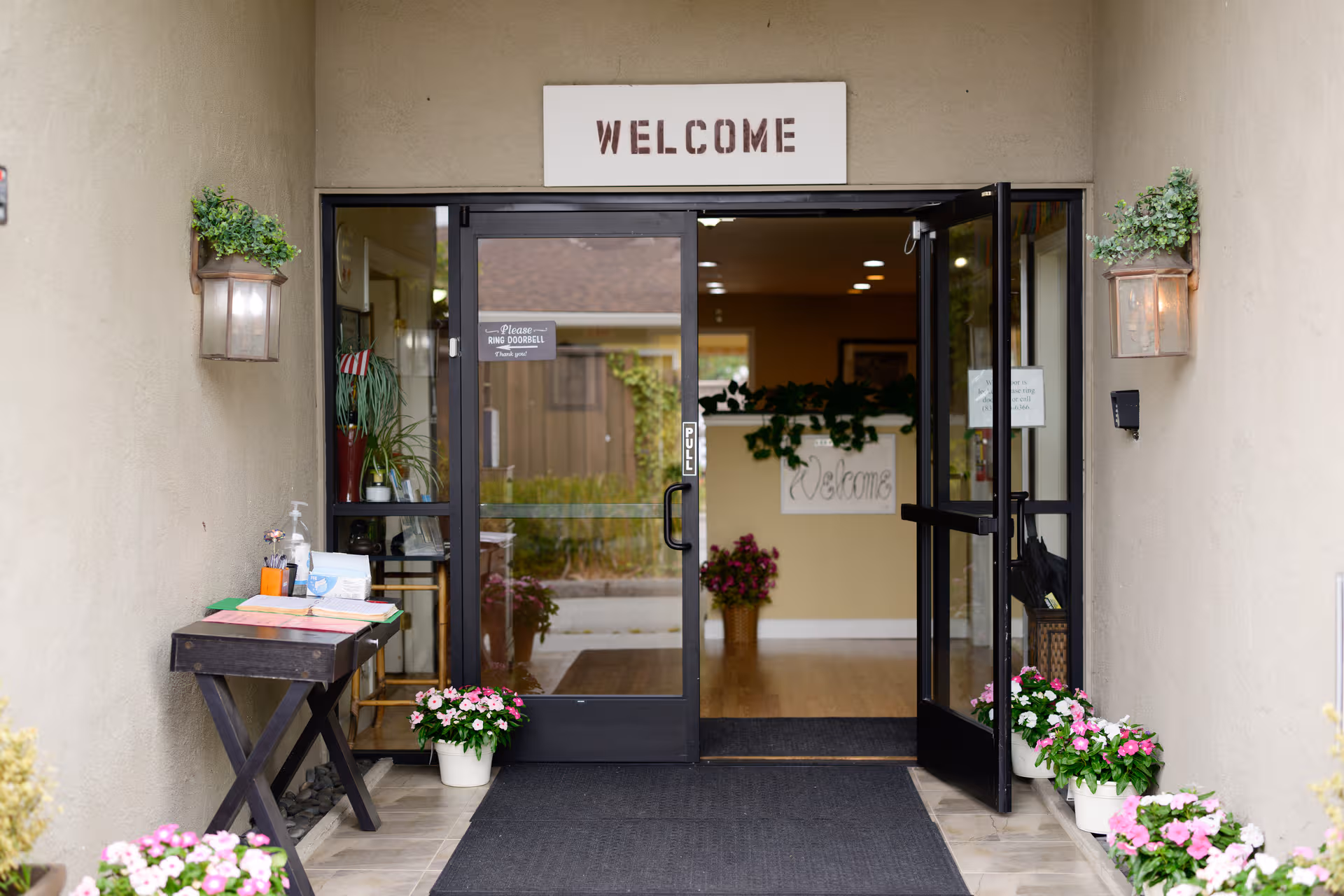 Open glass doors lead to a lobby with a Welcome sign and potted plants.