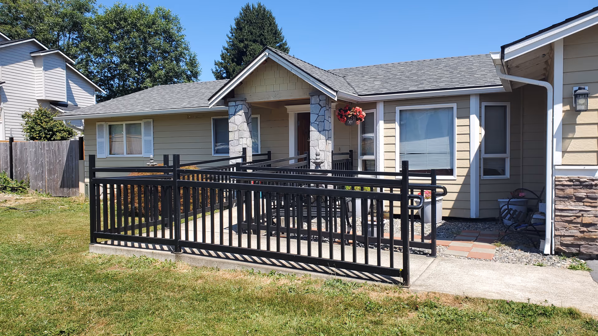 Front exterior view of a single-story beige house with a gray shingled roof, stone pillars at the entrance, a black metal wheelchair ramp, and a small garden area with grass and flowers under a clear blue sky.