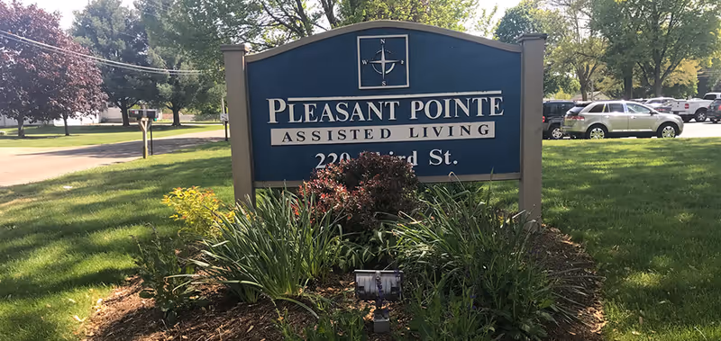 Blue entrance sign reading 'Pleasant Pointe Assisted Living' on a landscaped lawn with trees and parked cars in the background.