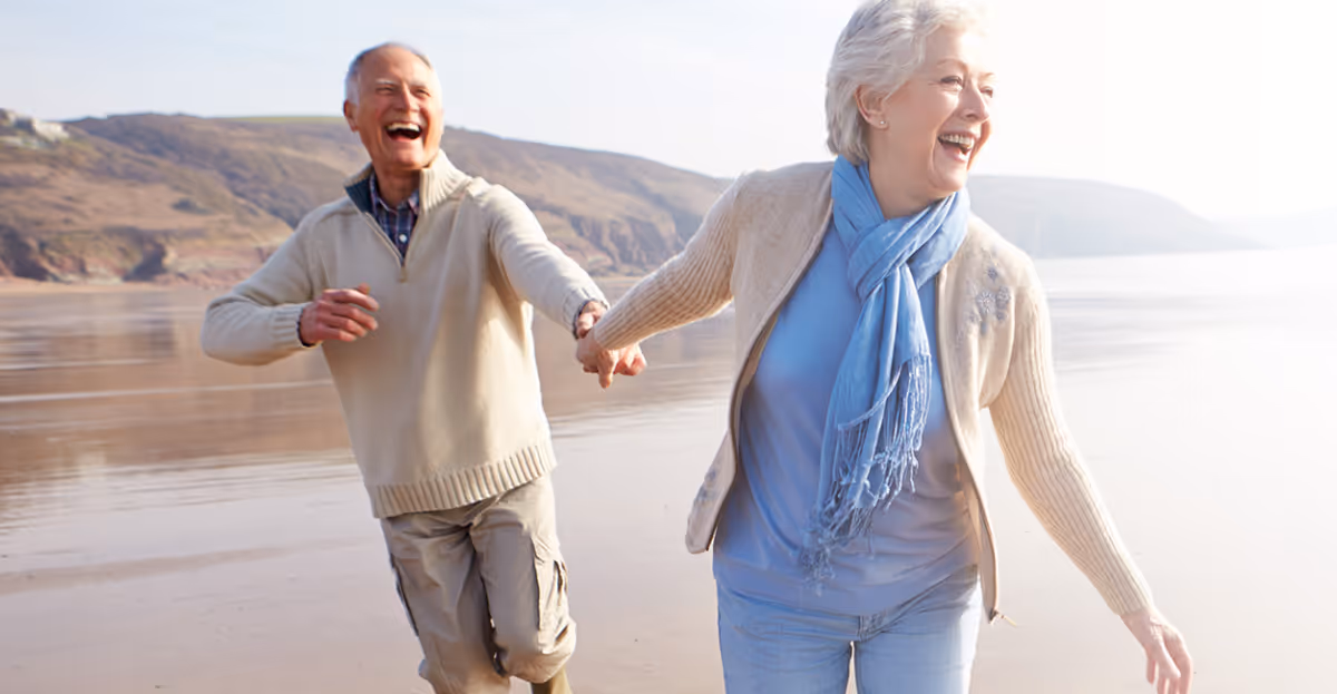 An elderly couple walks on a beach, holding hands and laughing.