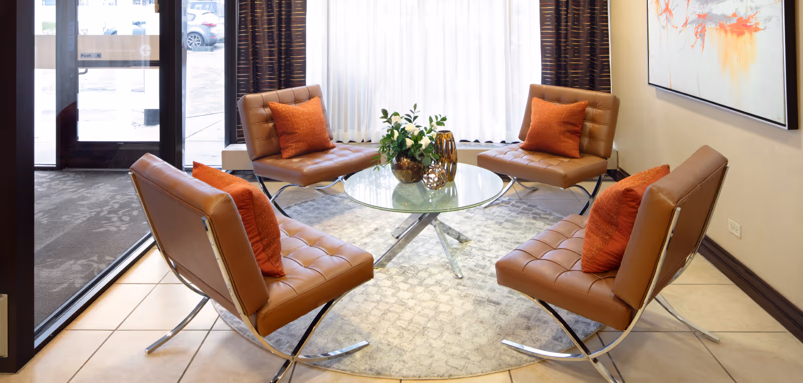 Four brown leather chairs with orange pillows arranged around a glass coffee table in a bright lounge near the entrance.