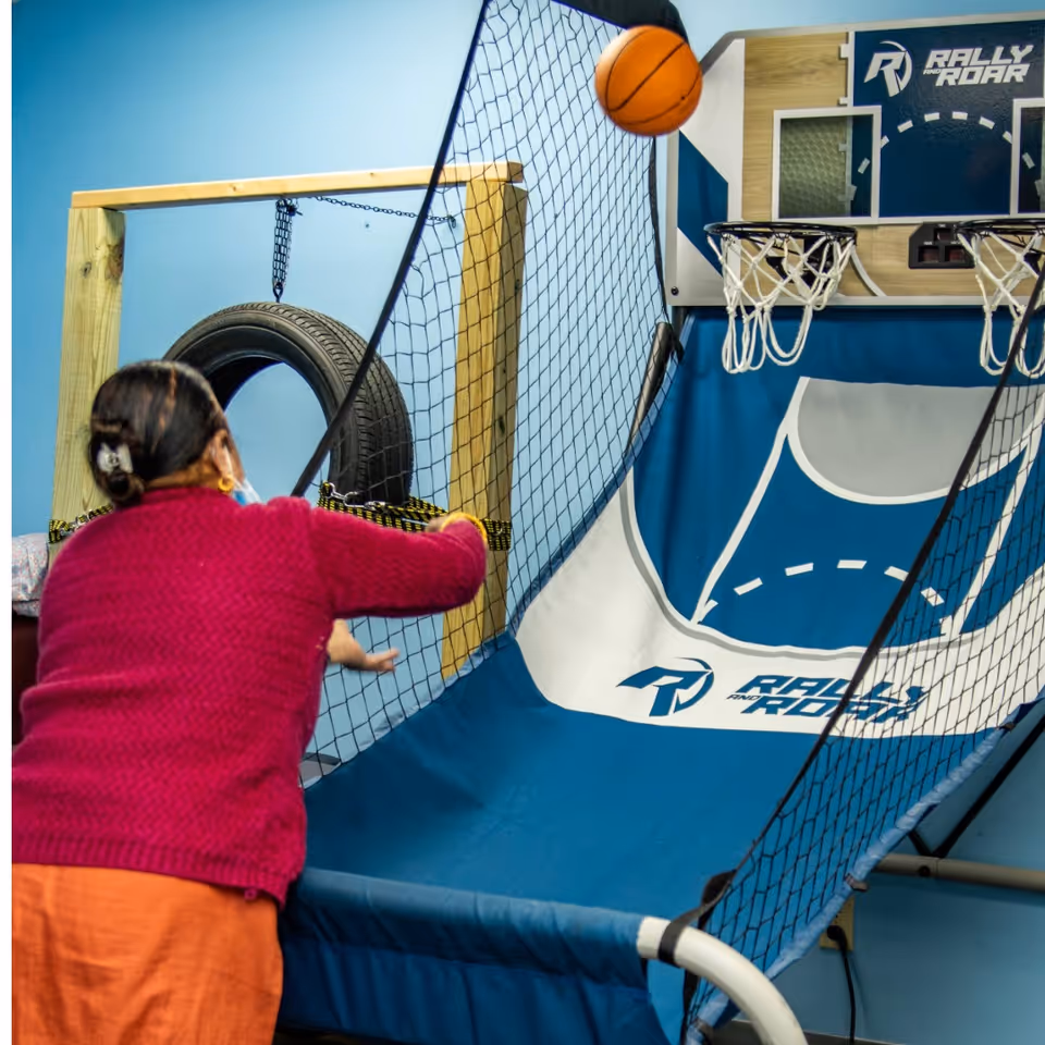 Resident in pink sweater engages in indoor athletic play area with netted basketball hoop.