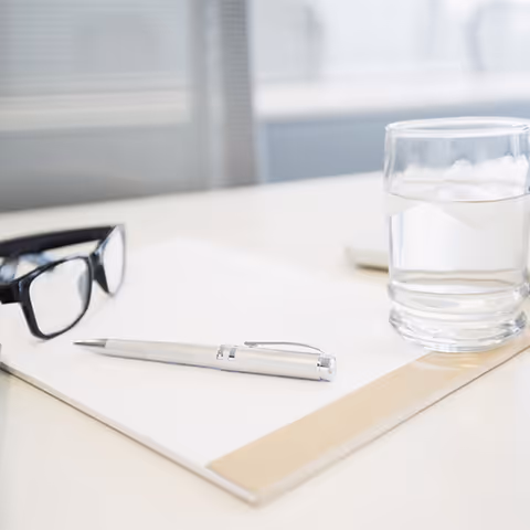 Close-up of a desk with glasses, a silver pen, paper, and a glass of water.