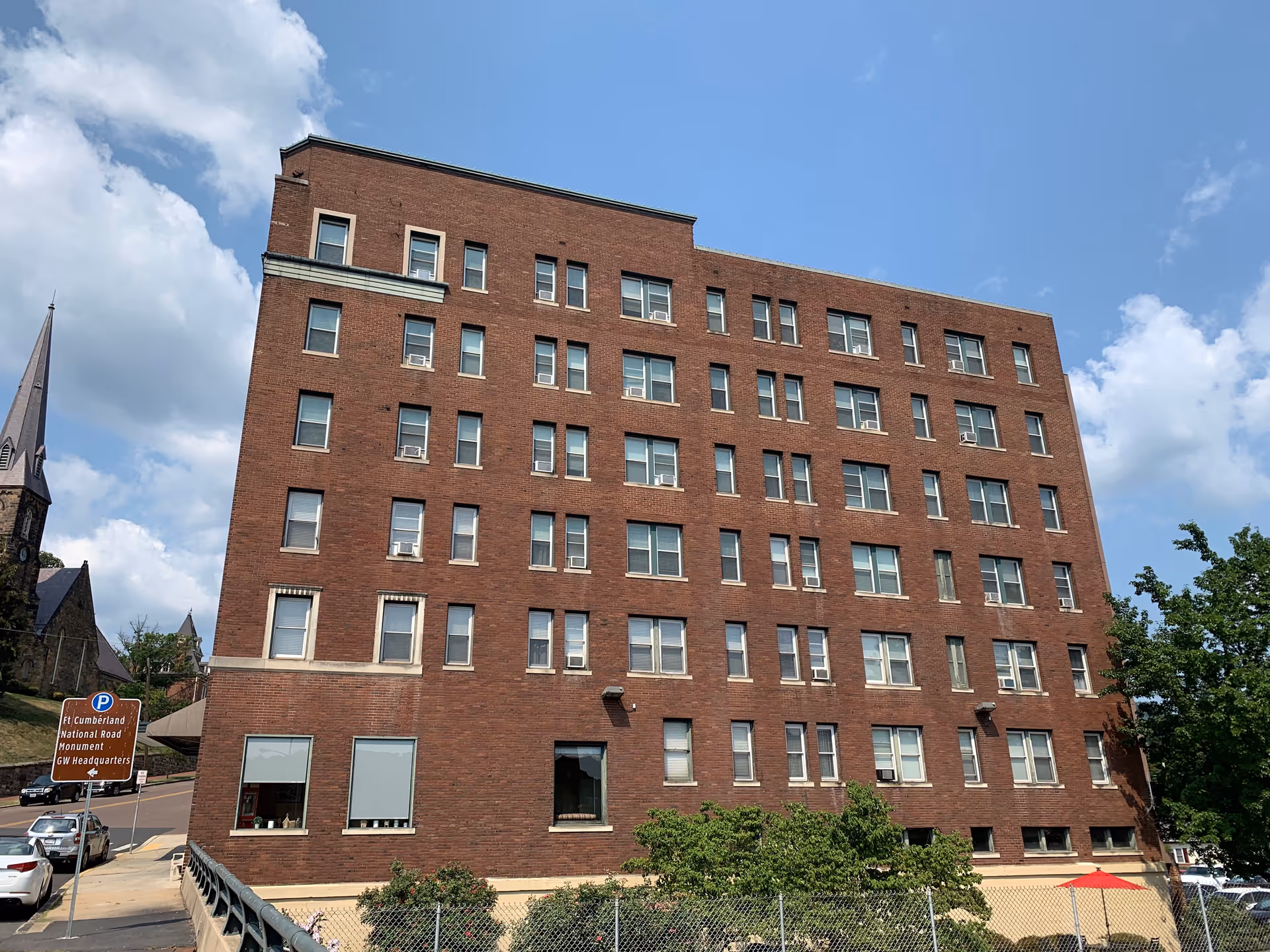 Front exterior of a red-brick, multi-story residential building seen from the street with a church steeple visible to the left.
