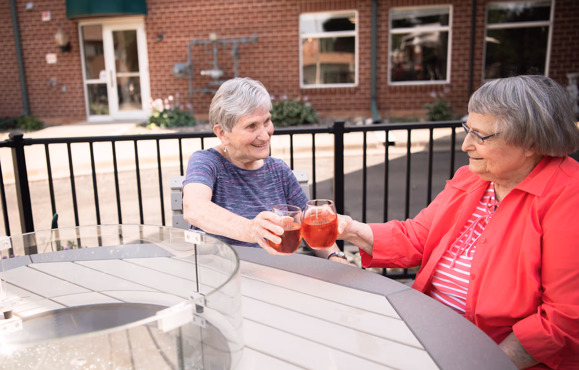 Two elderly women sitting at an outdoor table, smiling and clinking glasses with a reddish beverage. They are seated near a black metal fence with a brick building in the background.