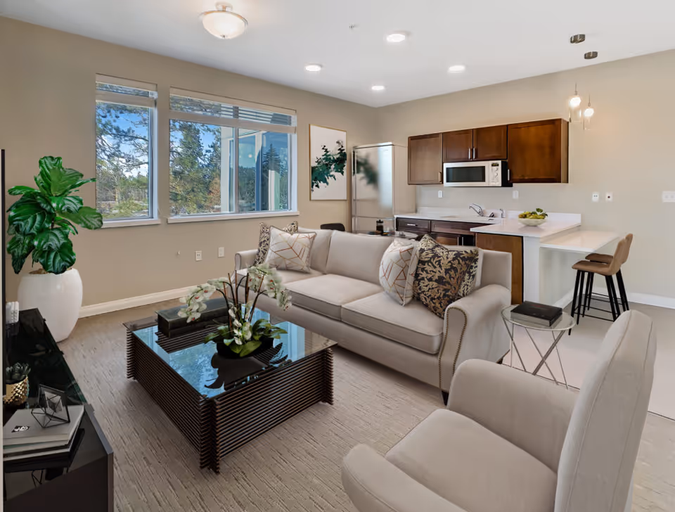 Bright open-plan living room with a beige sofa and coffee table facing a small kitchen with bar stools.
