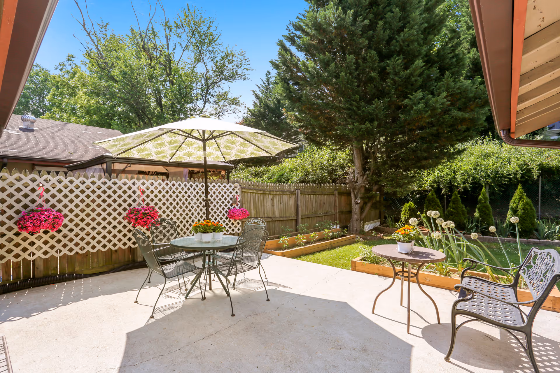 Outdoor patio area with a round glass table and four metal chairs under a large umbrella. There are hanging flower pots with pink flowers on a white lattice fence. The patio is surrounded by greenery including trees, bushes, and a small garden bed with plants. A metal bench and small round table with a flower pot are also visible on the patio.