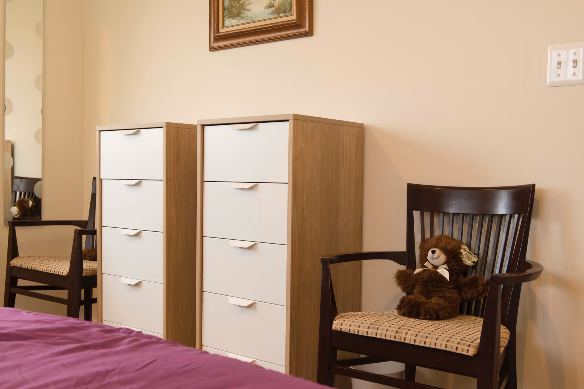 A bedroom corner with two wooden dressers featuring white drawers, a dark wooden chair with a patterned cushion, and a brown teddy bear sitting on the chair. A purple bedspread is partially visible in the foreground, and a framed painting hangs on the beige wall above the dressers.
