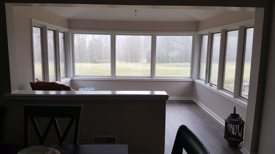 Interior sunroom-style living area with large wraparound windows overlooking a grassy yard.