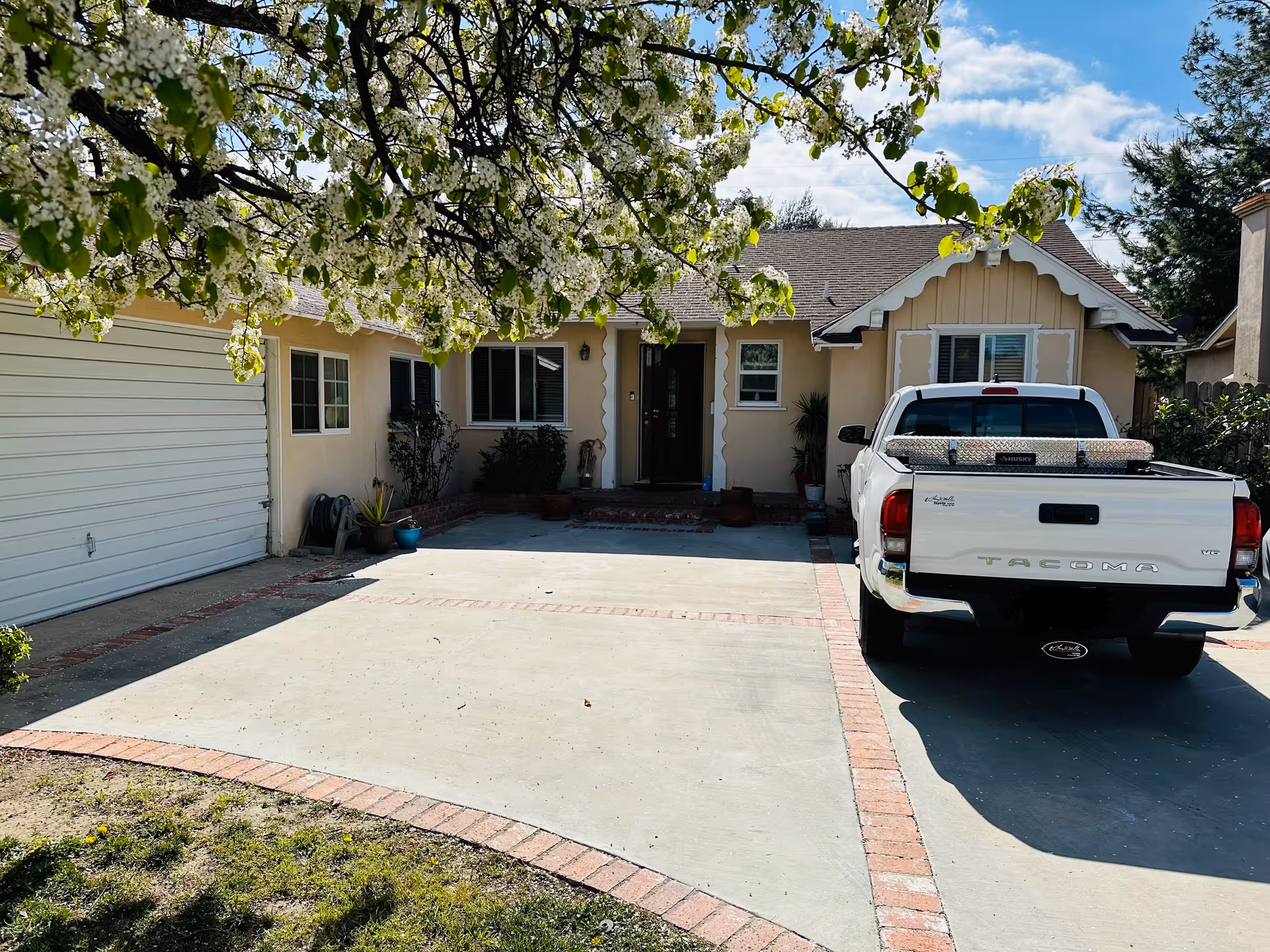 Front exterior of a single-story house with a garage, driveway, a parked white pickup truck, and flowering tree branches overhead.