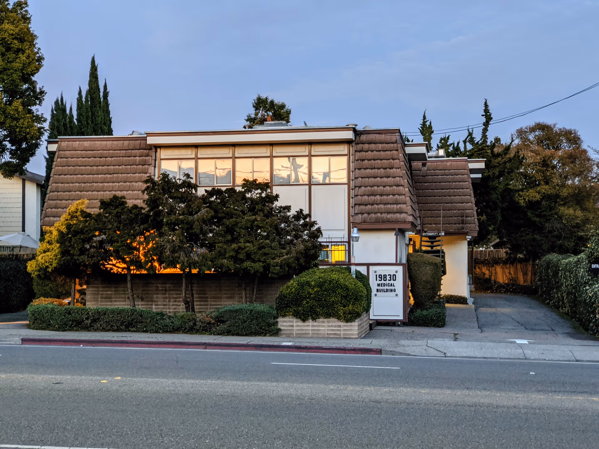 Front view of a brown, tiled-roof building with shrubs along a street.