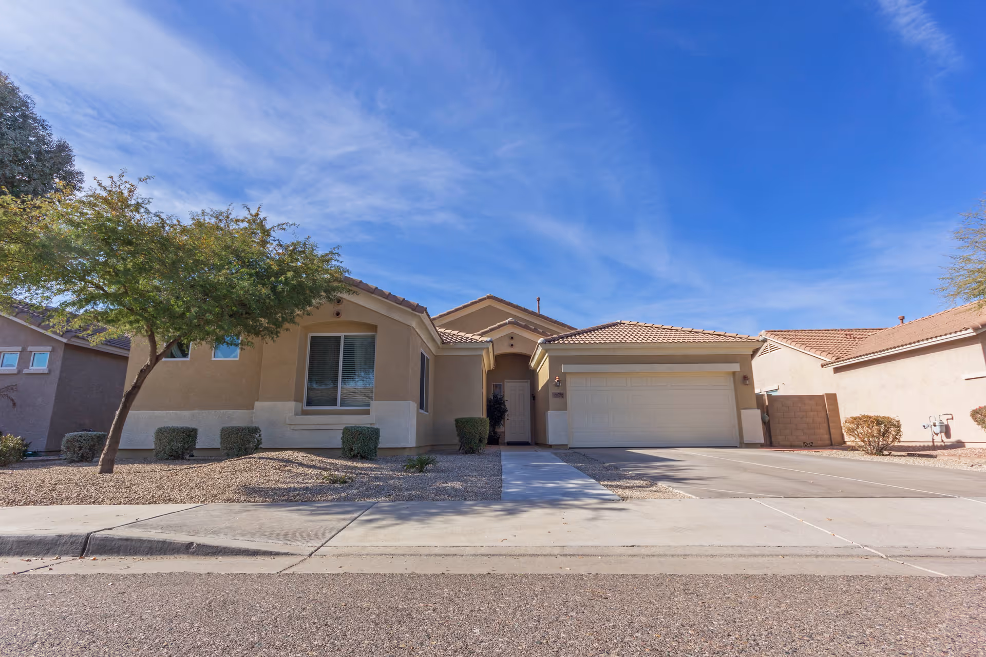 Single-story beige stucco house with a tiled roof, a two-car garage, a small tree, and desert landscaping in the front yard under a blue sky with some clouds.