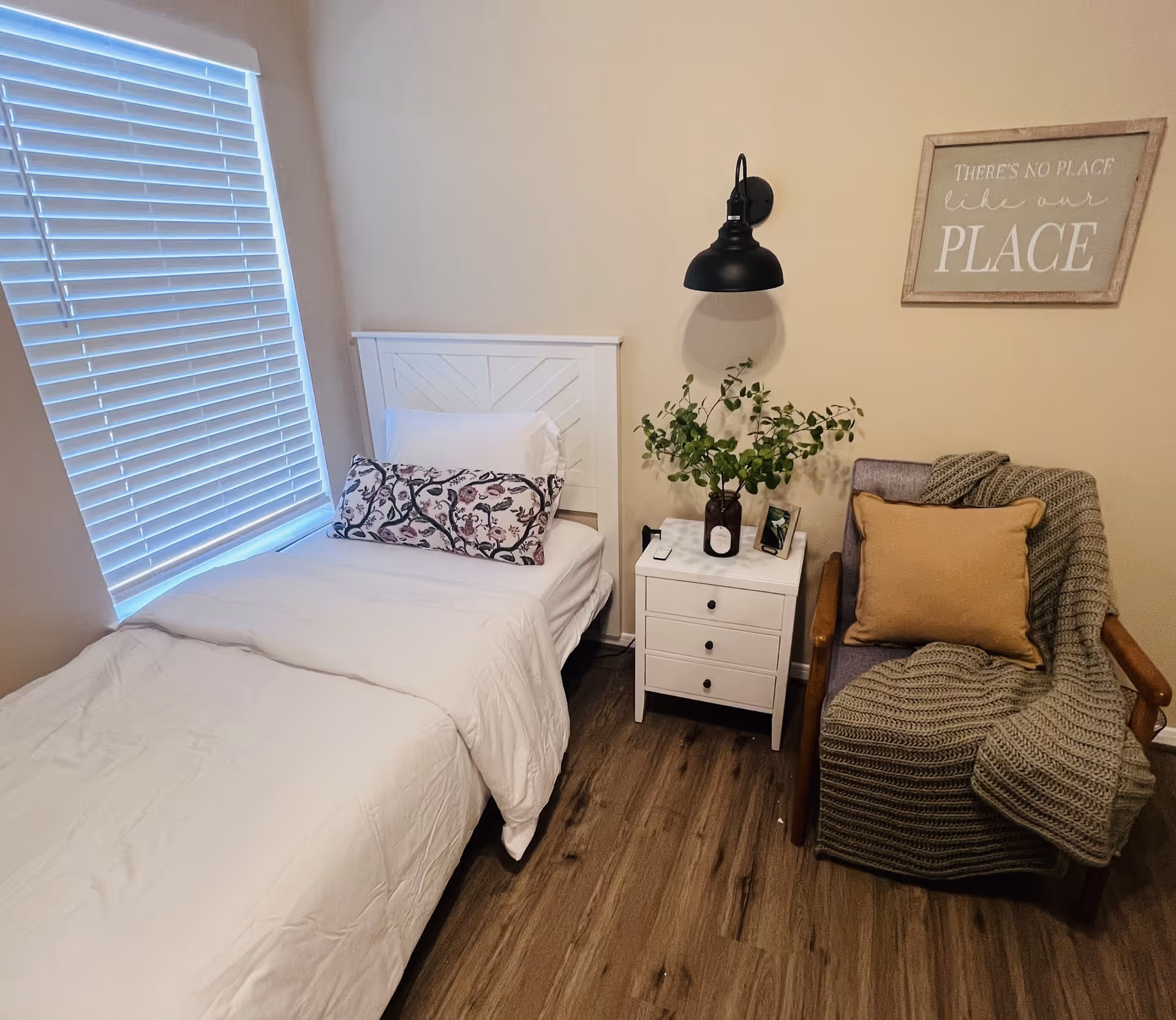 Neat small bedroom with a single bed, white bedside table topped with a plant and lamp, and an armchair draped with a throw and pillow.