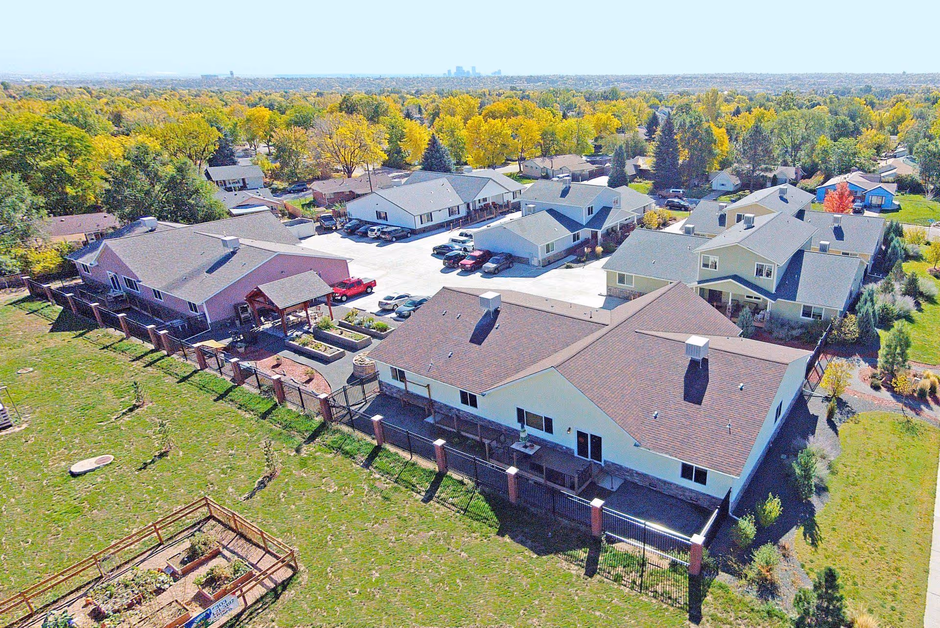 Aerial view of a senior living facility with multiple buildings, parking spaces with cars, surrounded by trees with autumn foliage and open grassy areas including a fenced garden plot.