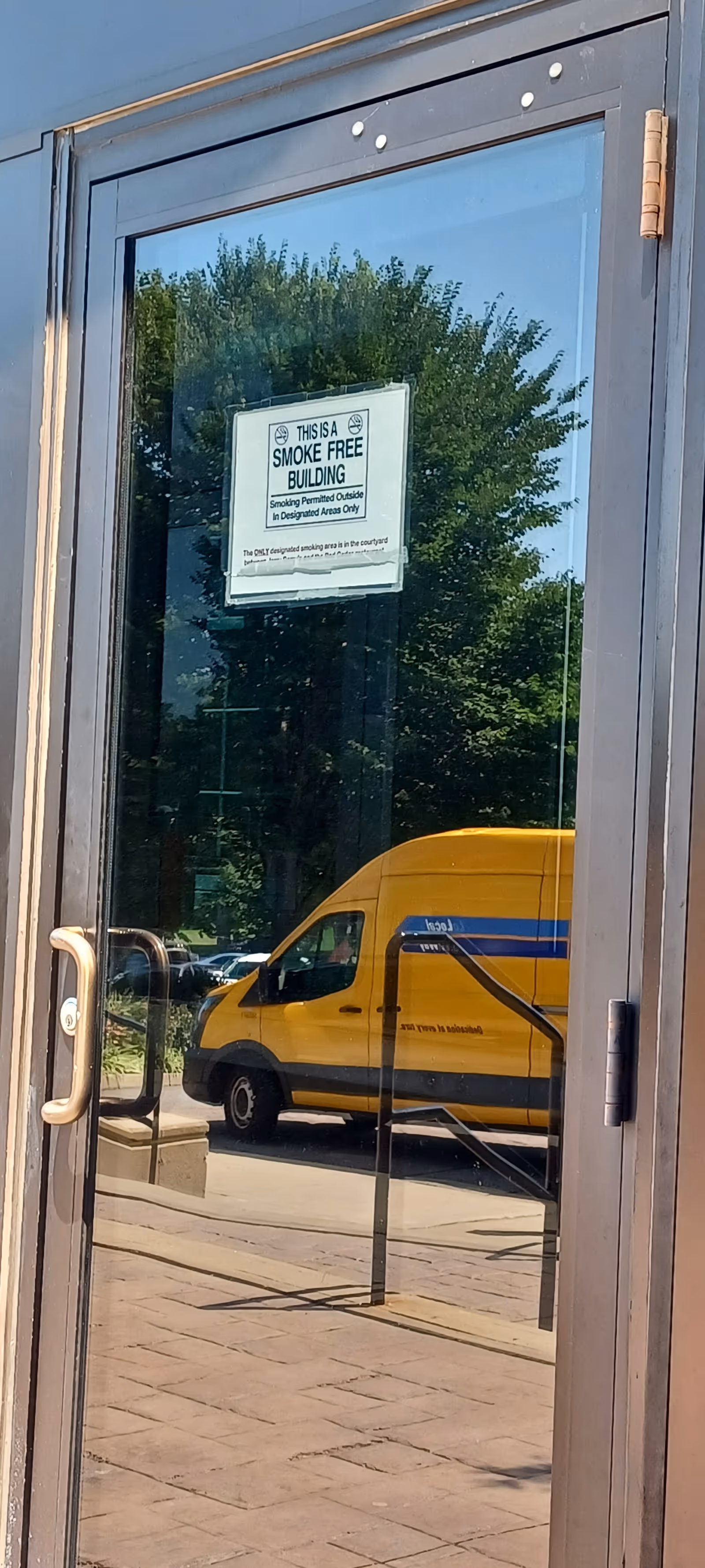 Glass door reflecting trees and a yellow van, with a smoke-free building sign attached.