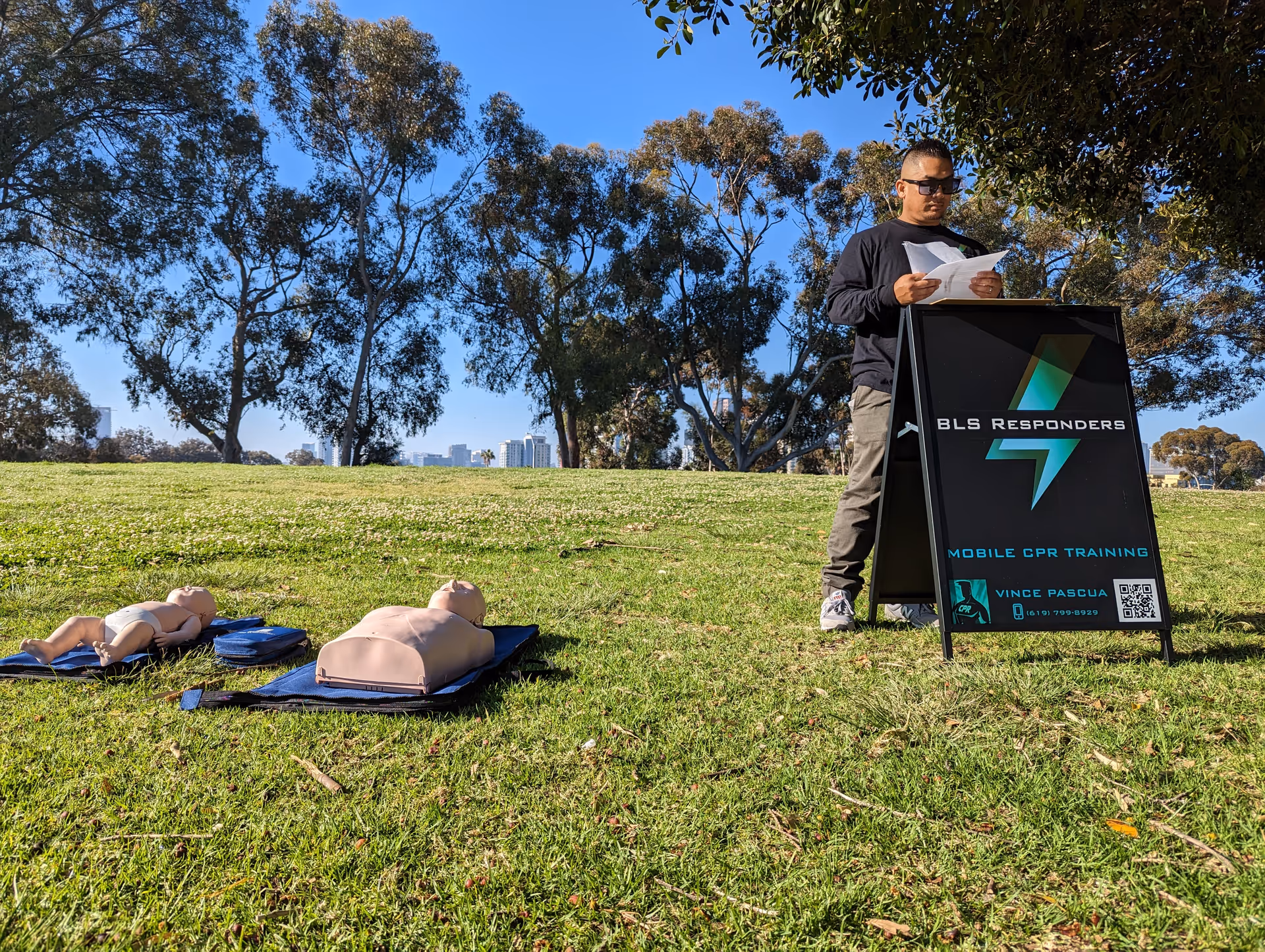 Trainer reads papers beside BLS Responders CPR sign with outdoor mannequins on grass.