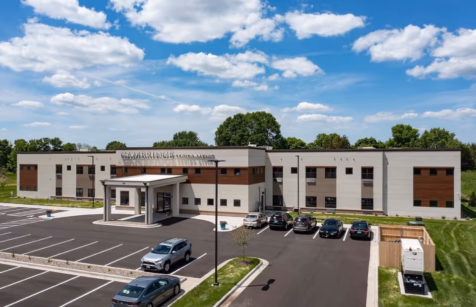 A modern two story senior living facility with a covered entrance, parking lot, and green surroundings.