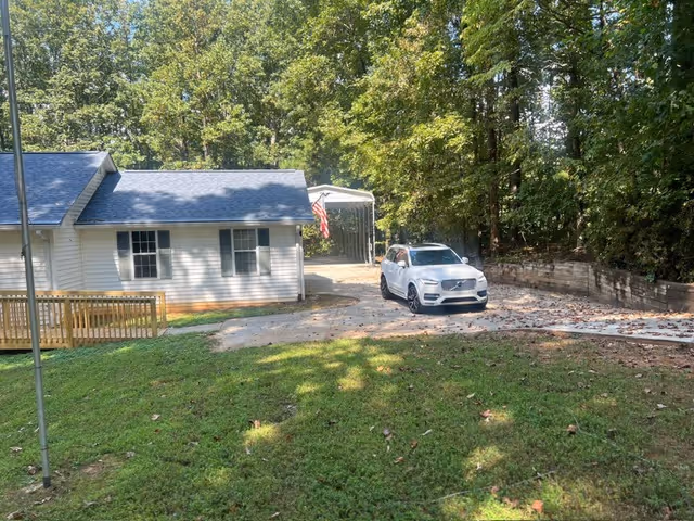 A single-story care facility exterior with a driveway, white car, and surrounding trees.