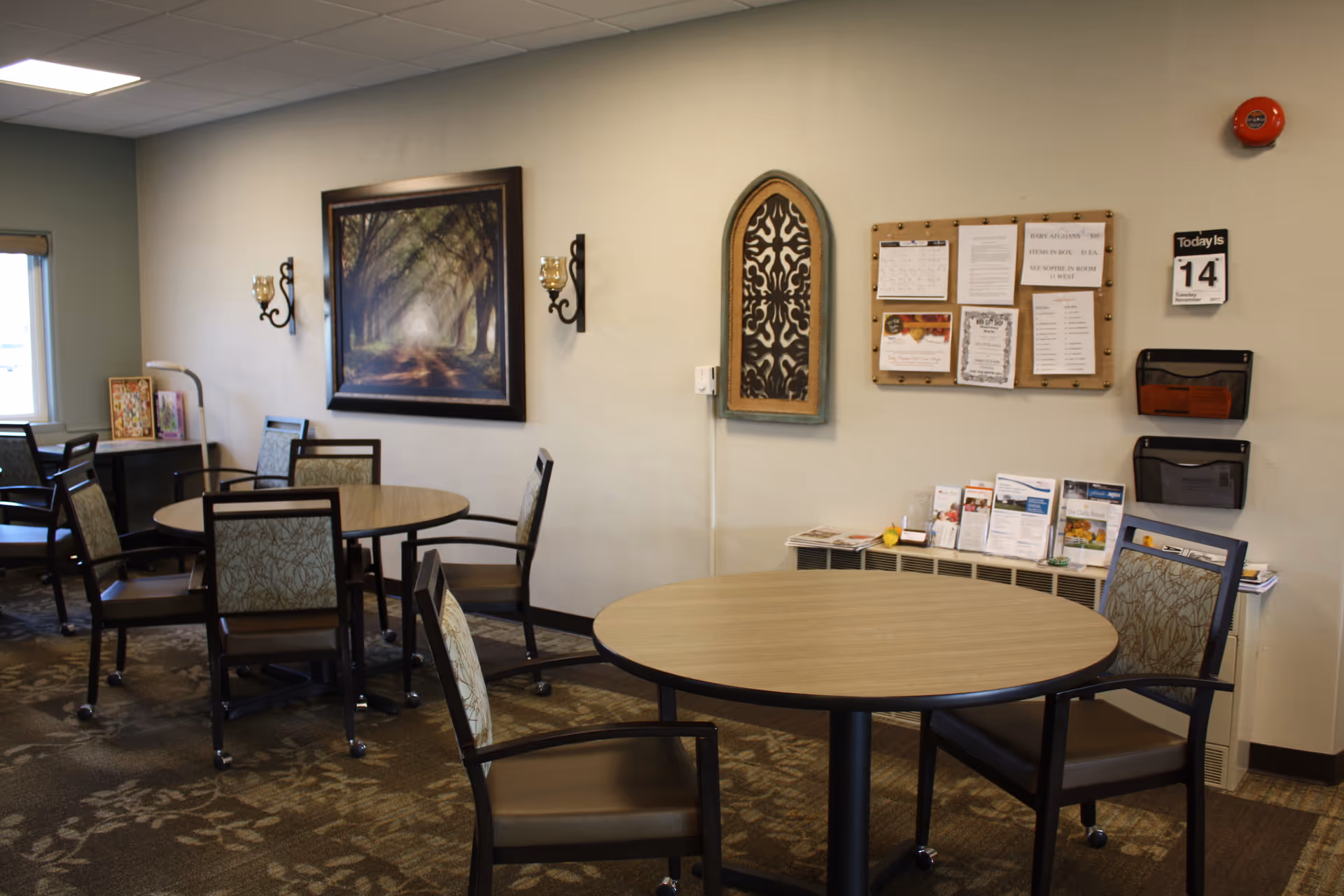 Senior living facility common area with round tables, chairs, wall art, and a bulletin board.