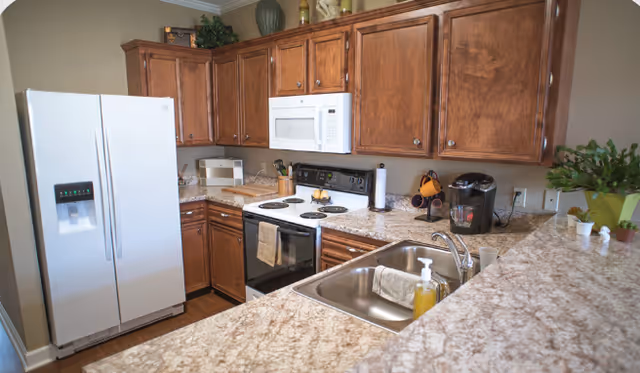 A kitchen with brown wooden cabinets, appliances, countertops, and a sink in a senior living unit.
