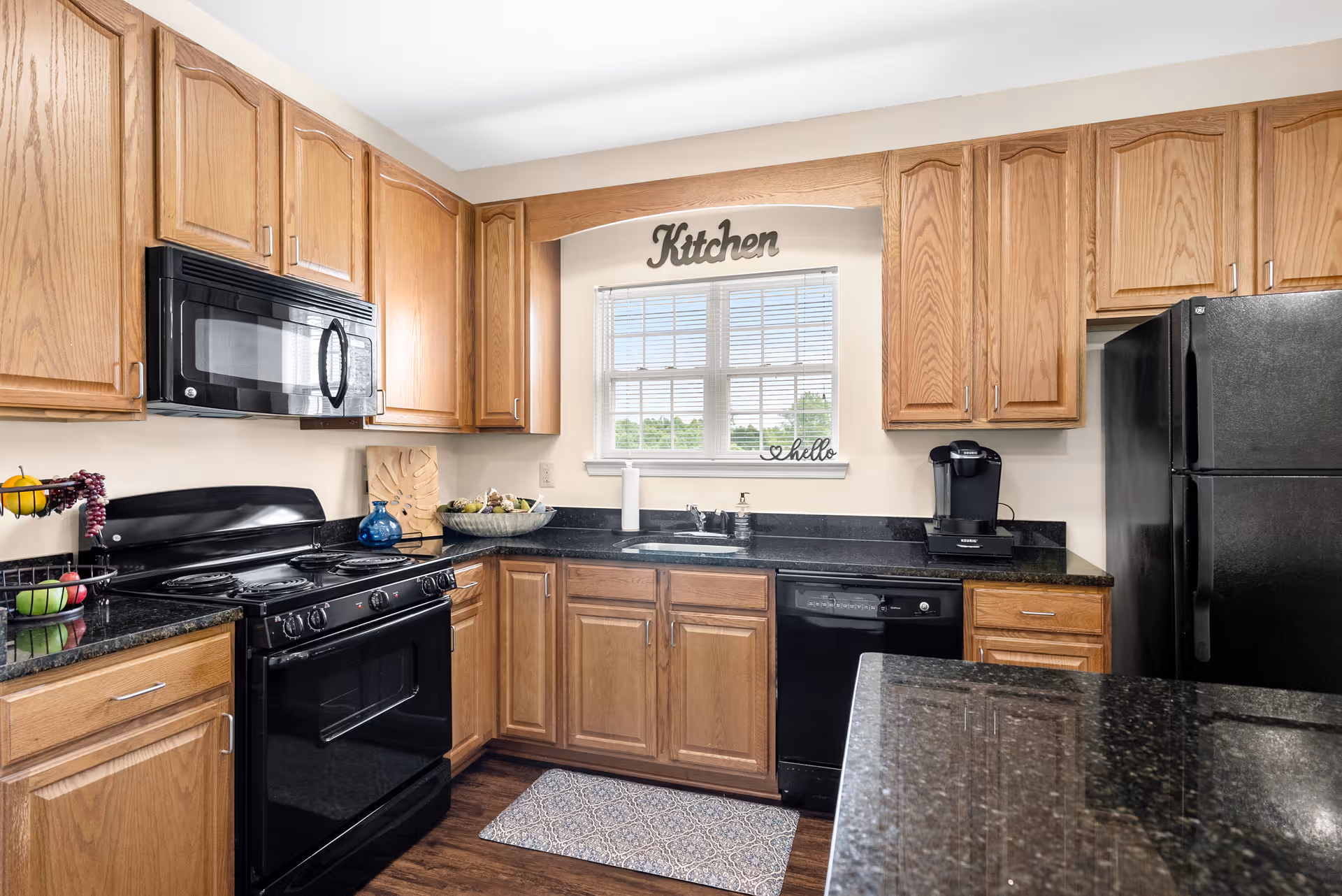 A bright kitchen with wooden cabinets, black appliances including a stove, microwave, dishwasher, and refrigerator. There is a window above the sink with a decorative sign that says 'Kitchen' and a small 'hello' sign on the windowsill. The countertops are black granite, and there is a small rug on the wooden floor in front of the sink.