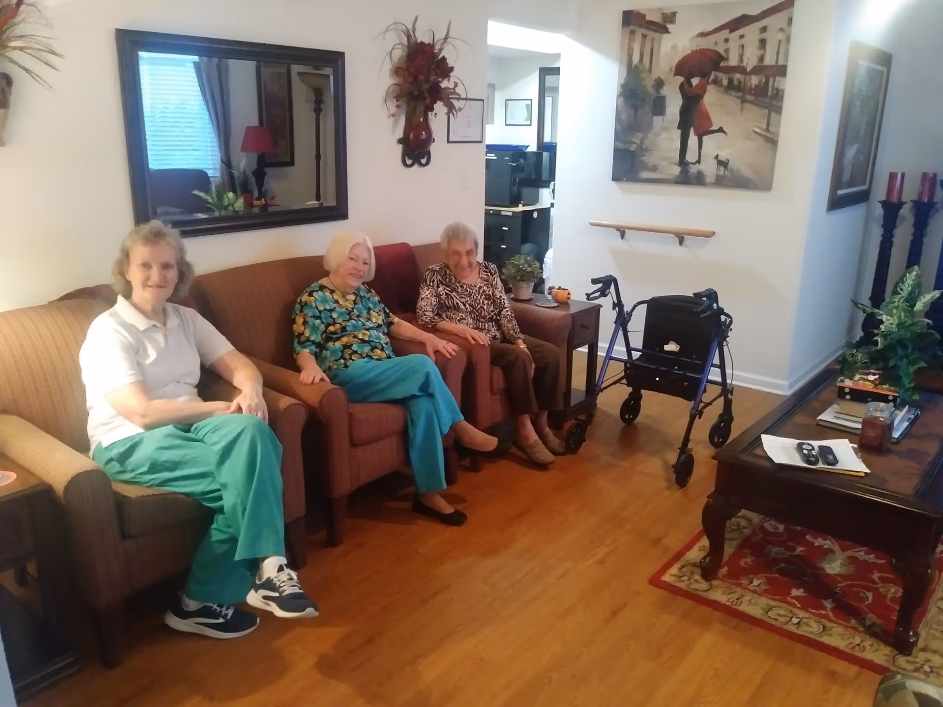 Three elderly women sitting on couches in a cozy living room with wooden flooring. There is a walker next to one of the women, a coffee table with remote controls and decorative items, a large mirror on the wall, and framed artwork. The room is warmly decorated with plants and candles.