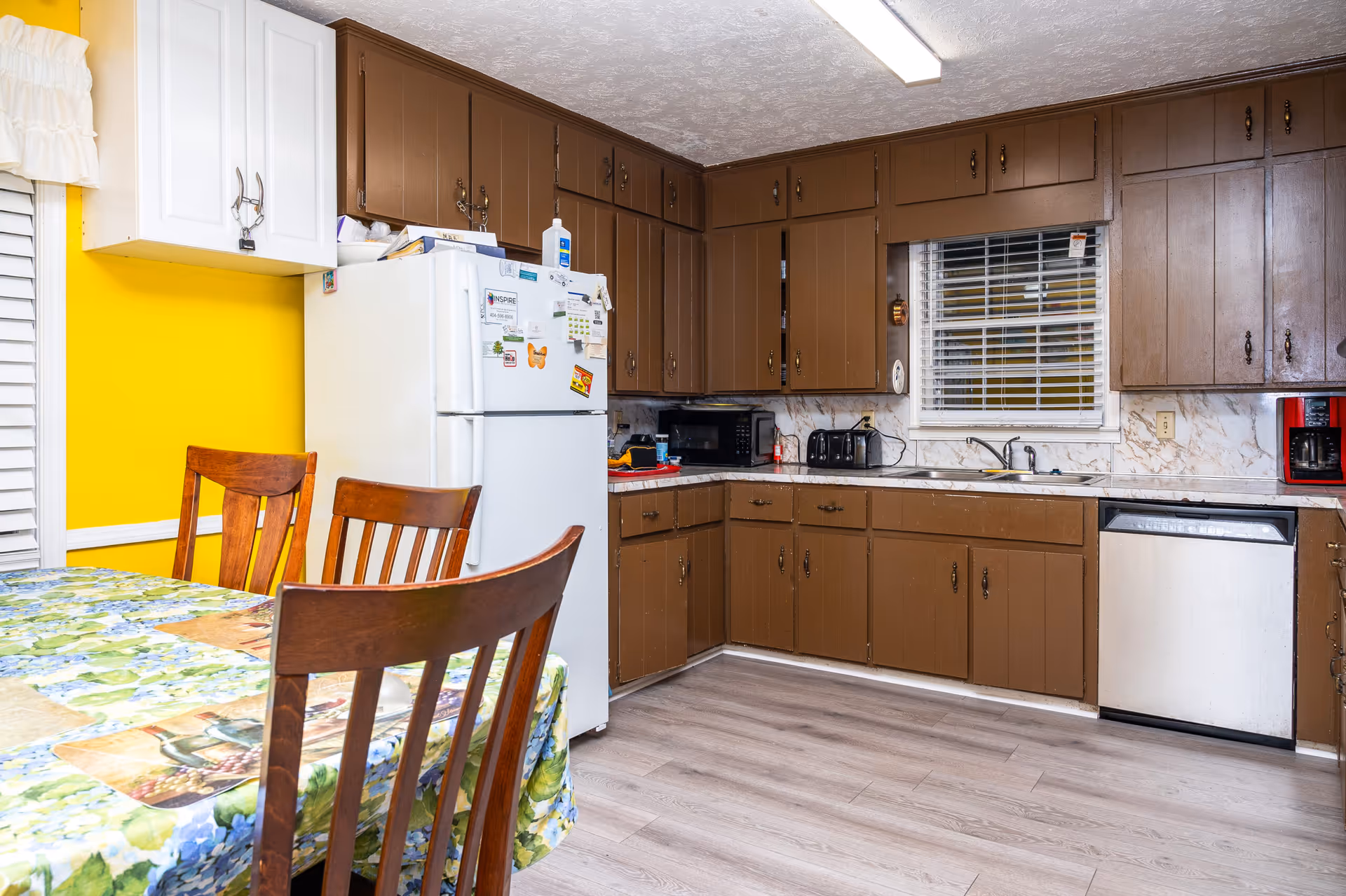 Brown kitchen cabinets surround a white refrigerator and table in a bright kitchen.