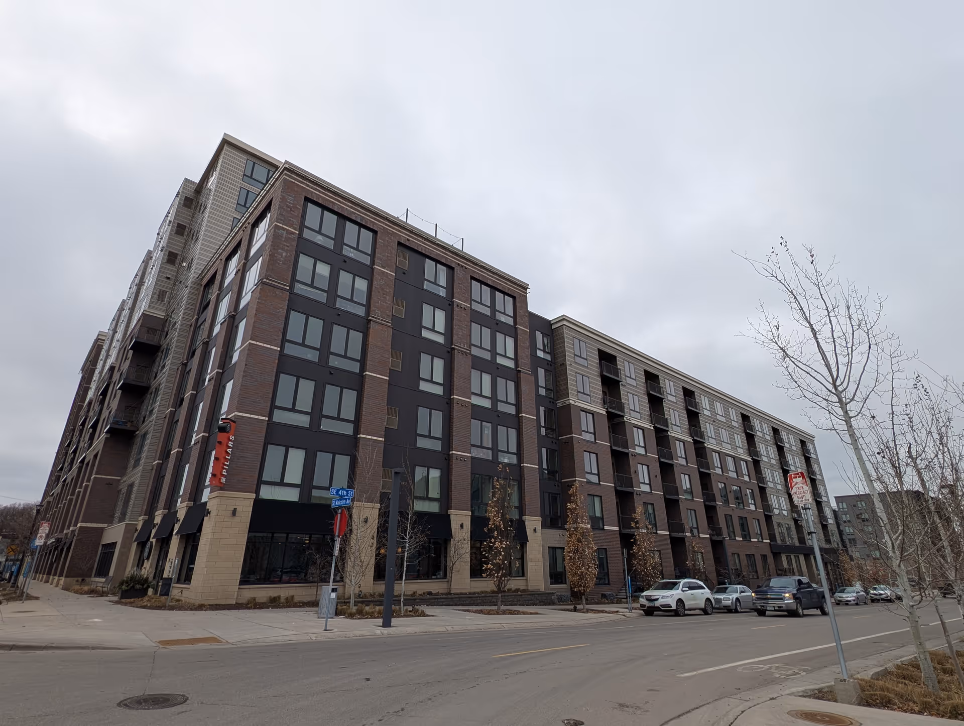 Street-level view of a multi-story brick and glass residential building with parked cars and bare trees under an overcast sky.