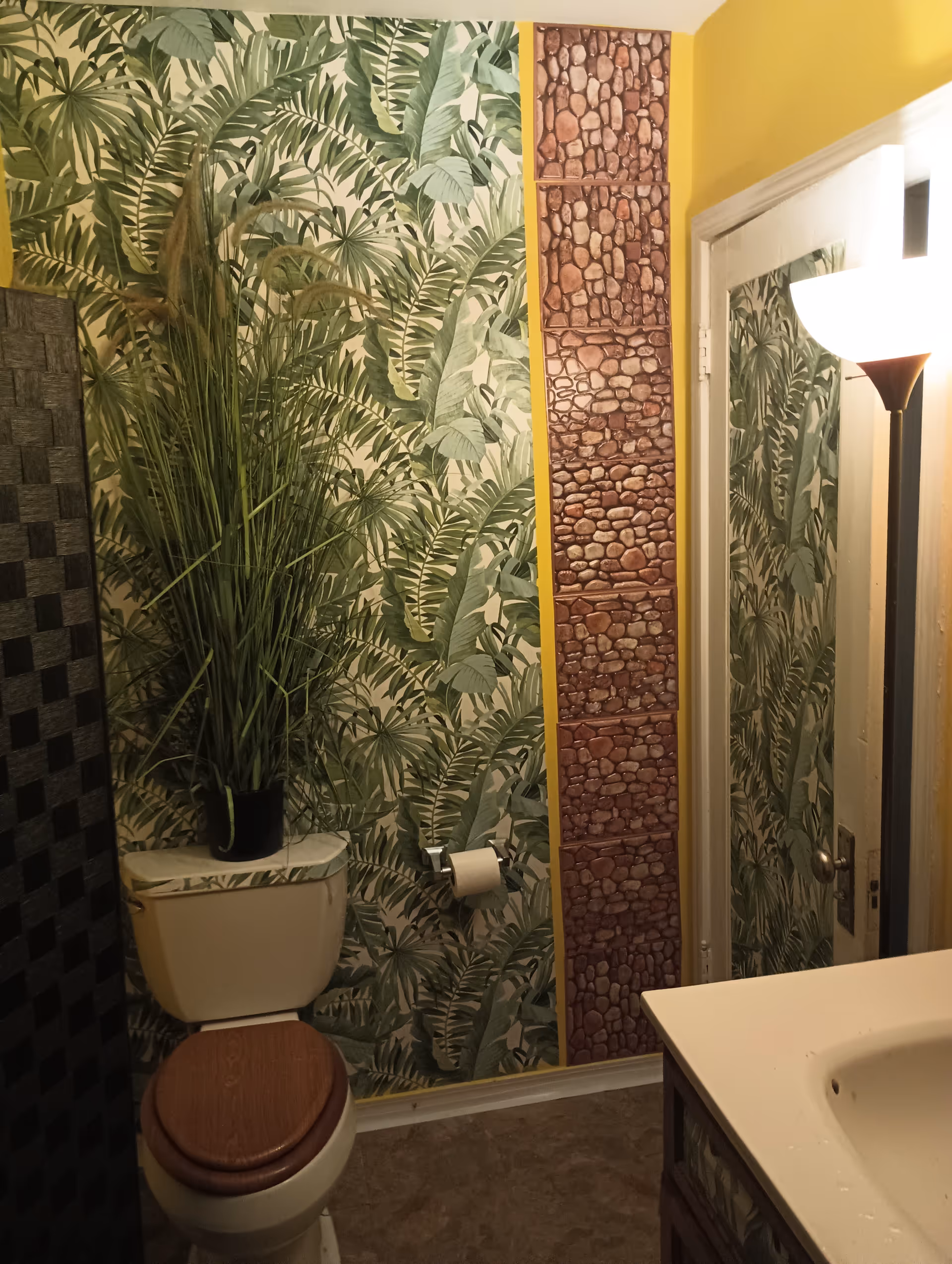 Tropical plant-themed bathroom with toilet, sink, mirror, and stone-pattern wall panel.