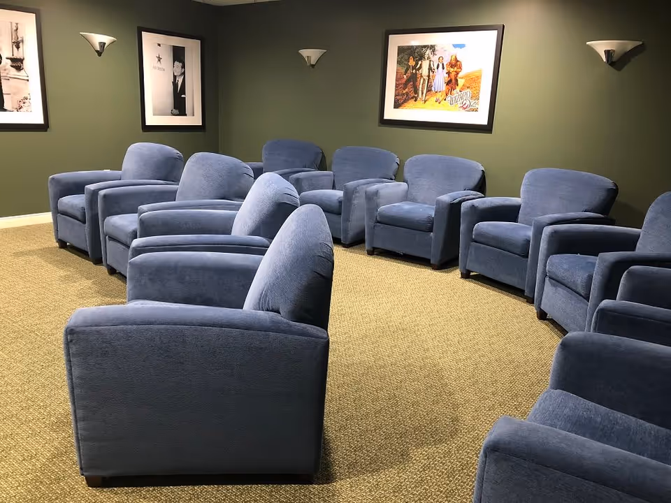 A room with multiple blue upholstered armchairs arranged in a semi-circle on a carpeted floor. The walls are painted green and decorated with framed pictures, including a colorful poster of The Wizard of Oz. Wall-mounted light fixtures provide illumination.