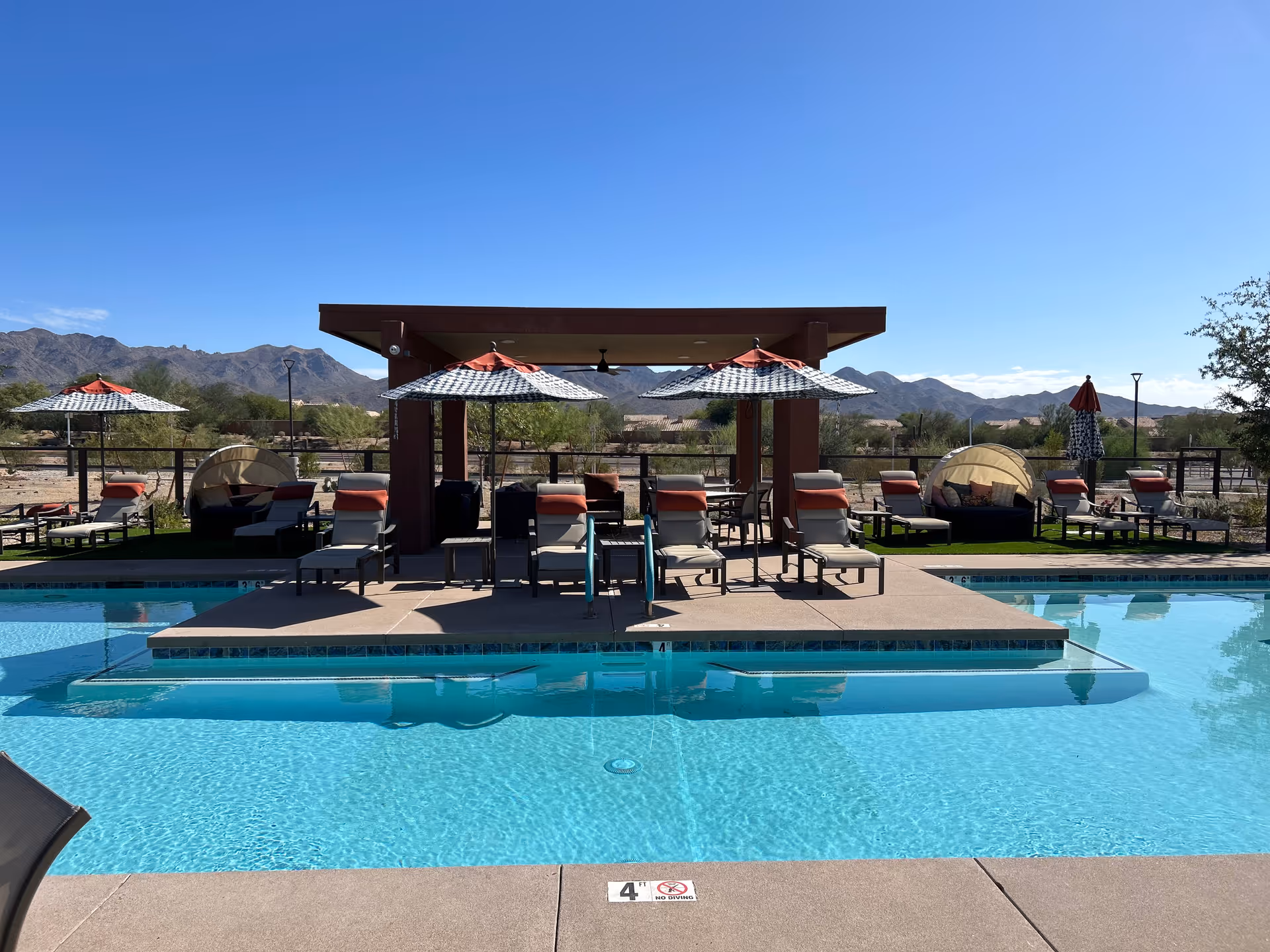 Outdoor pool area with lounge chairs, umbrellas, cabanas, and desert mountains in background.