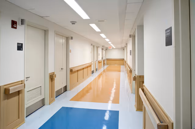 Long, brightly lit interior hallway in a nursing facility with handrails and colored linoleum flooring.