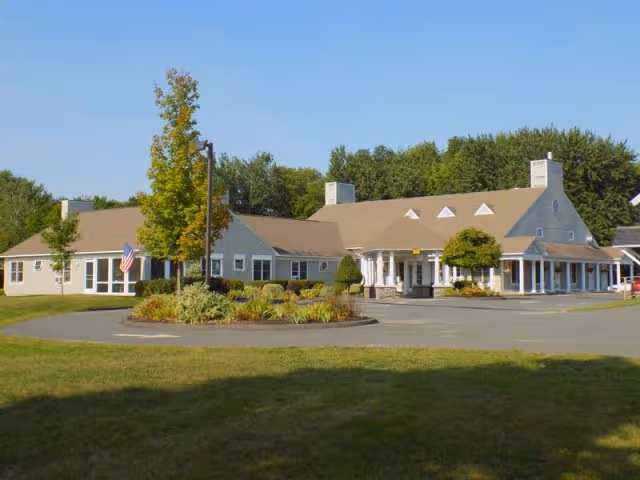 Front exterior of a single-story facility with a covered entrance, circular driveway, landscaped island, and an American flag.
