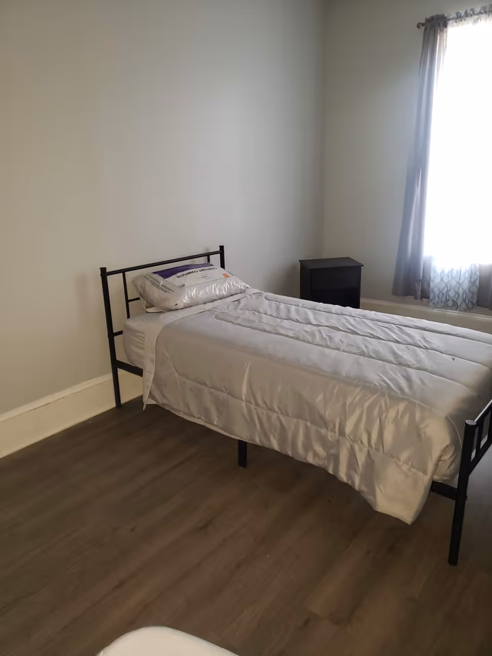 A small bedroom with a single bed, nightstand, and window with curtains.