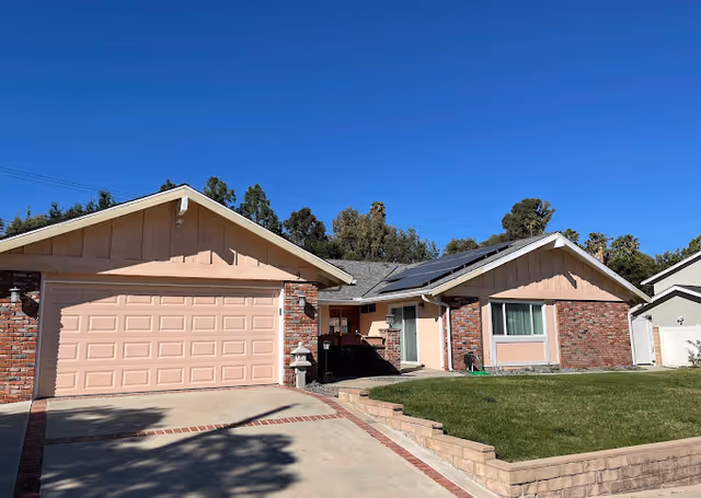 Single-story ranch-style house with an attached two-car garage, brick and siding facade, solar panels on the roof, and a front lawn under a clear blue sky.