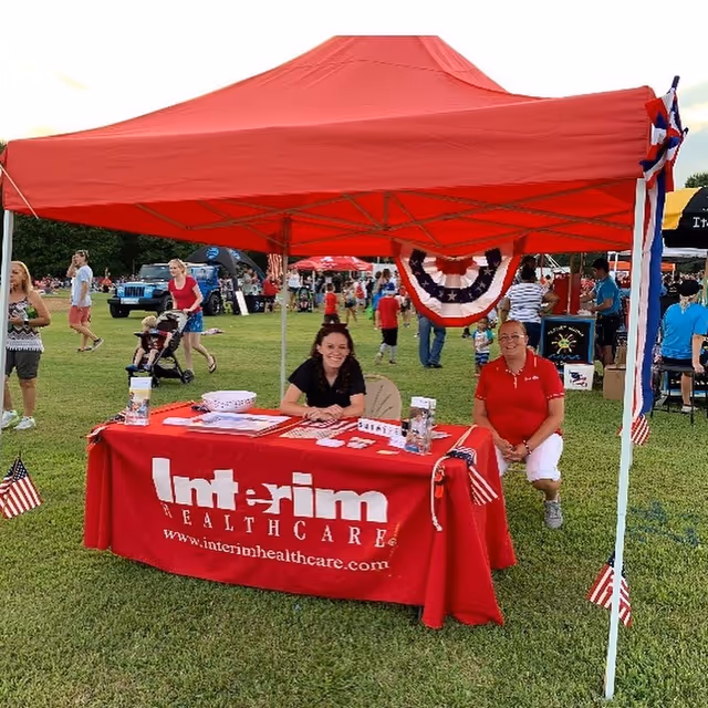 Two people sit at an outdoor Interim HealthCare booth with a red table on grass.