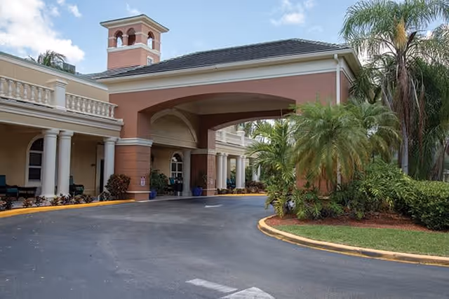 Exterior view of Brookdale West Boynton Beach facility entrance with a covered driveway, columns, palm trees, and landscaped greenery under a partly cloudy sky.