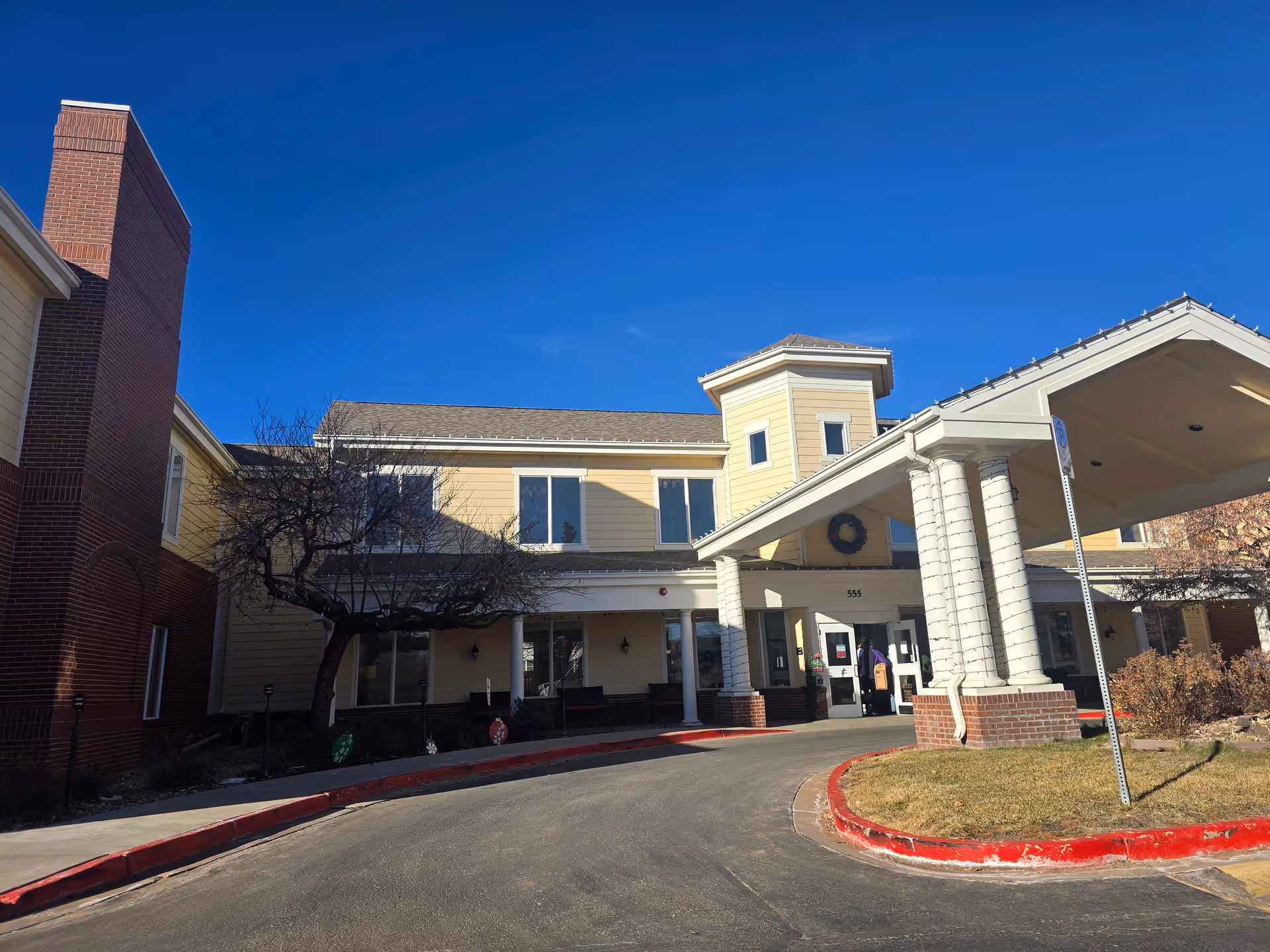 Front entrance and porte-cochère of a two-story assisted living building with columns, a driveway, and a clear blue sky.