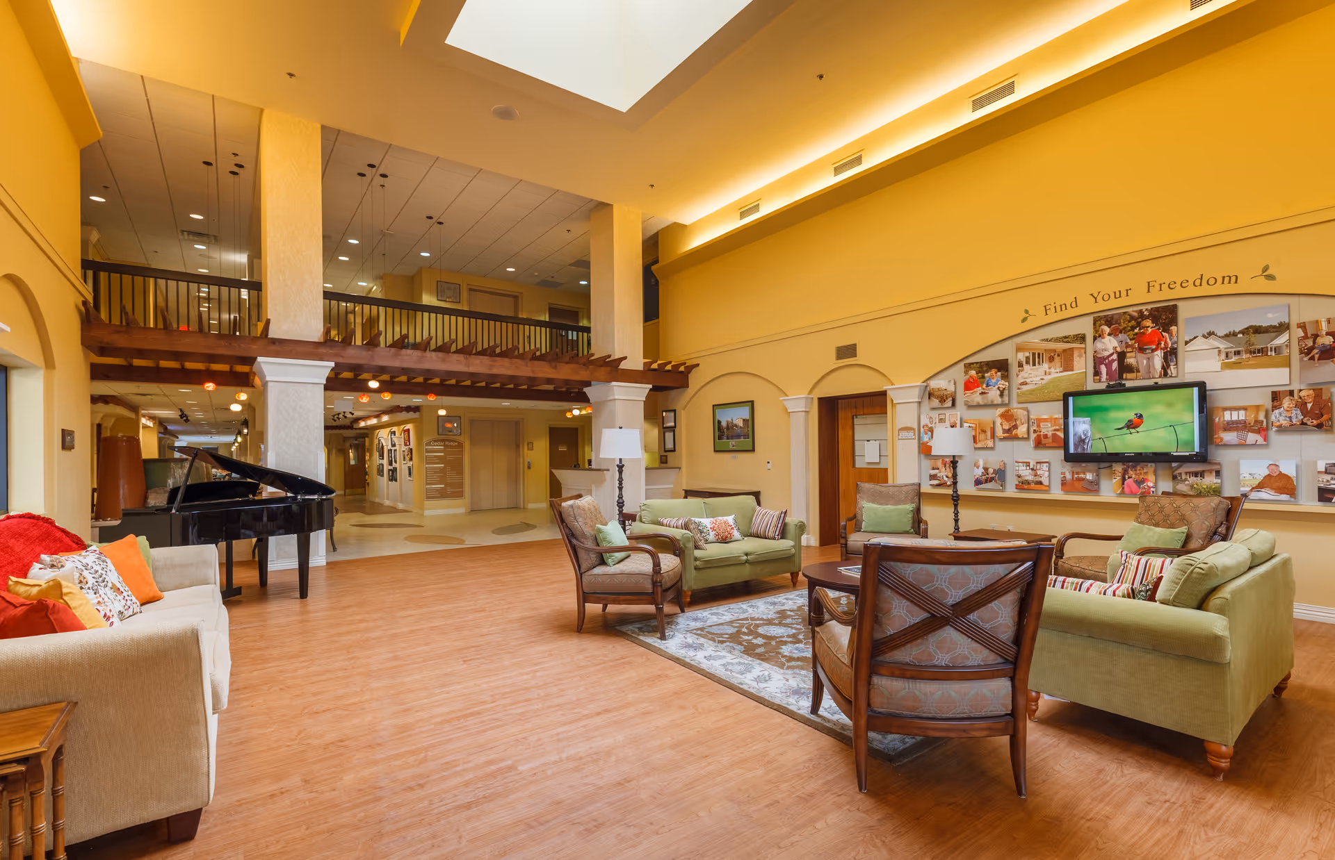 A spacious and well-lit common area in a senior living facility with yellow walls and wooden flooring. The room features comfortable seating including green sofas and armchairs arranged around a coffee table on a patterned rug. A grand piano is positioned near the left side of the room. The back wall displays a collage of photos under the phrase 'Find Your Freedom' with a flat-screen TV showing a bird. The area has a high ceiling with a skylight and an upper balcony overlooking the space.
