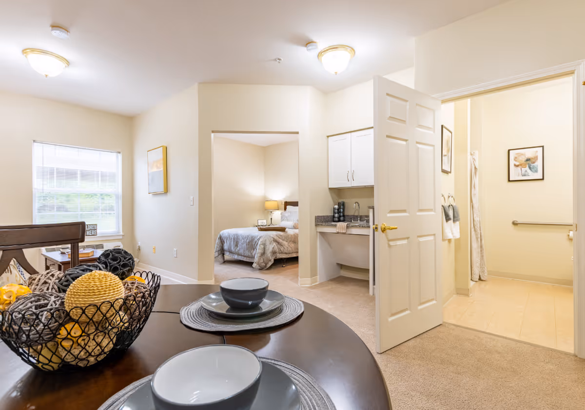 Interior dining area with a round table, adjacent bedroom, and kitchenette in a senior living facility.