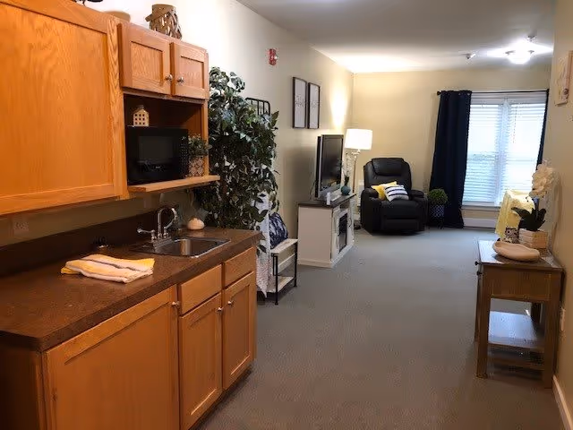 Interior view of a senior living facility room showing a small kitchenette with wooden cabinets and a sink on the left, a living area with a black recliner chair, a TV on a white stand, a lamp, and a window with dark curtains in the background. There is a small table with decorative items on the right side of the room.