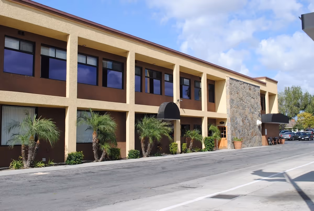Two-story tan building with brown trim, palm trees, a stone wall, and a parking lot.