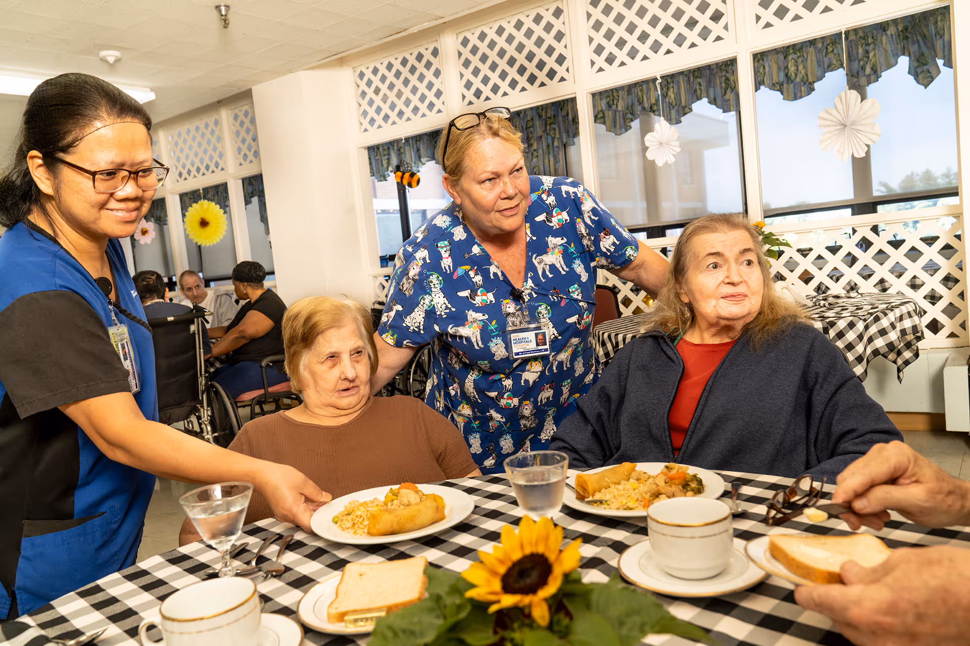 Two elderly women seated at a table with checkered tablecloths in a dining area, being served food by a staff member in blue scrubs while another staff member in patterned scrubs stands behind them. The table has plates of food, cups, glasses of water, and a sunflower centerpiece.