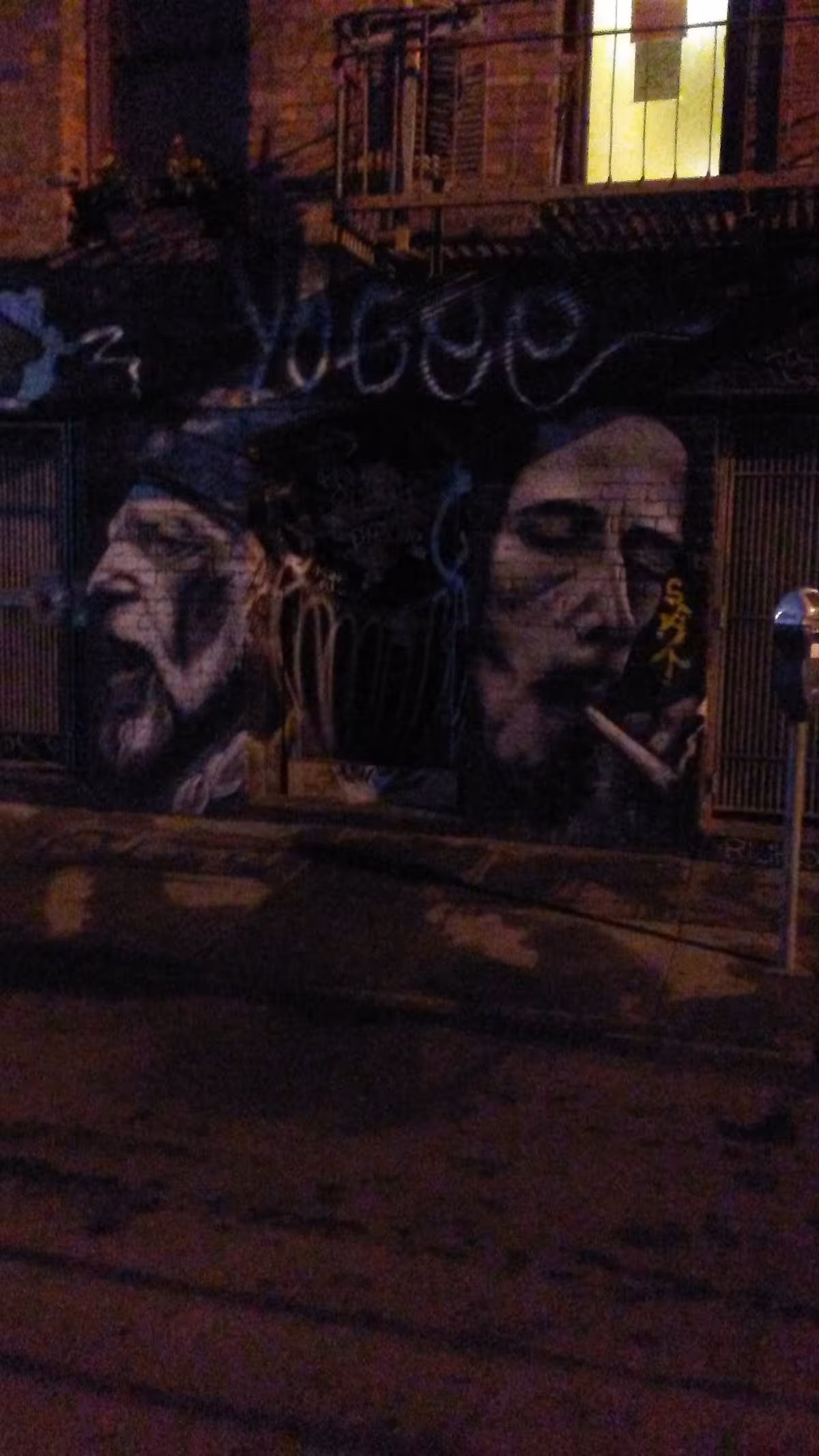 Dimly lit exterior wall featuring large graffiti portraits and a lit upstairs window.