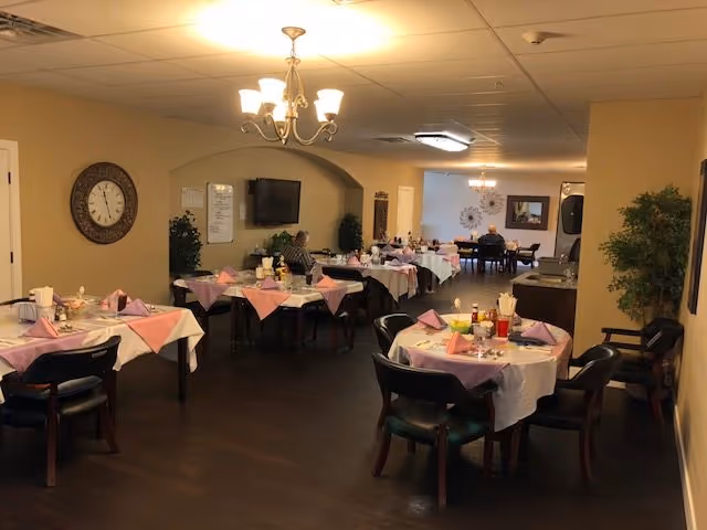Dining room with multiple round tables set with pink napkins and chairs, chandeliers, a wall clock, a TV, and potted plants.