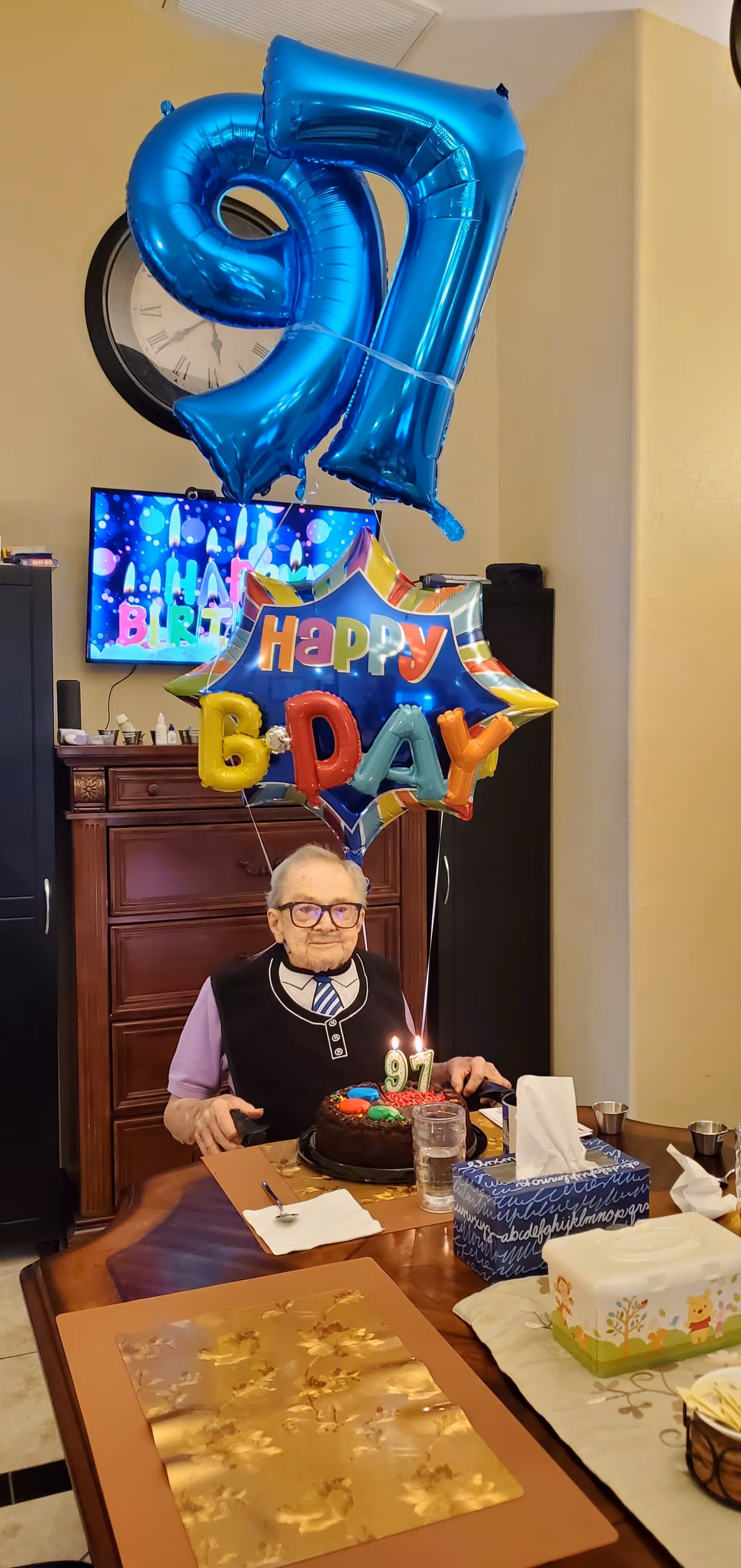 An elderly man sitting at a dining table with a birthday cake in front of him. The cake has lit candles shaped as the number 97. Behind him are large blue balloons shaped as the number 97 and a colorful star-shaped balloon with the words 'Happy B Day'. A television screen in the background displays a birthday-themed image. The setting appears to be a cozy indoor dining area with wooden furniture.
